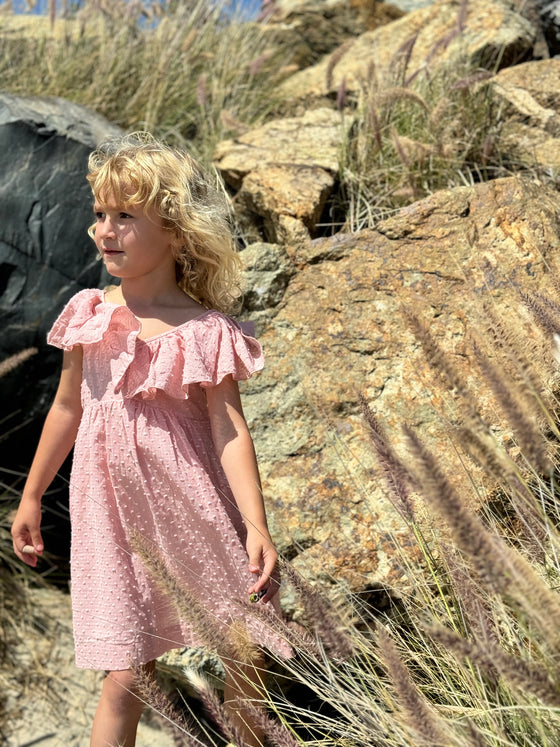 a little girl looks off into the distance while wearing a pink Swiss dot sun dress with ruffled collar. 