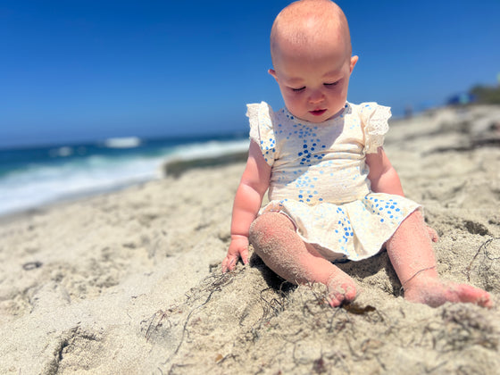 A baby sitting on the beach wearing a white and blue floral print skirted bloomer with matching onesie in the same print. 