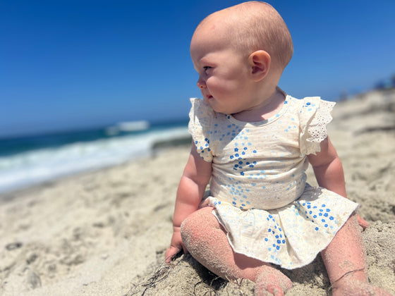 A bay sitting on the beach looking off into the distance while wearing a matching white and blue skirted bloomer with matching onesie