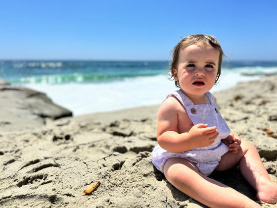 A baby on the beach wearing a purple baby embroidered pinafore bubble 