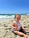 A brunette baby at the beach wearing a purple overall onesie bubble 