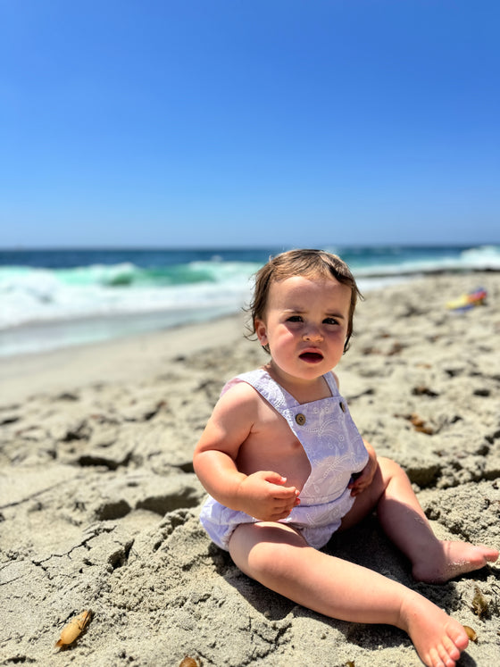 A brunette baby at the beach wearing a purple overall onesie bubble 