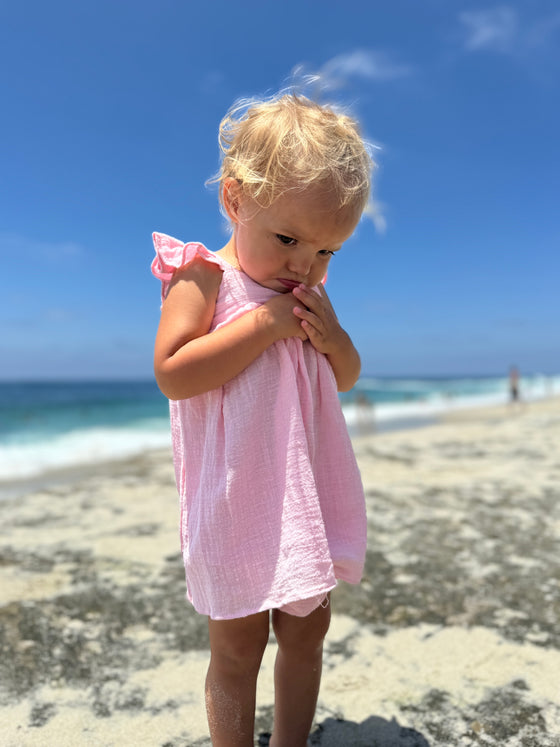 Girl toddler looking down while  playing on the beach in pink dress with ruffled sleeves 