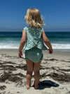 Child model on the beach wearing Green Plaid Aria dress with matching bloomer showing back view with riffle criss-cross detail.  