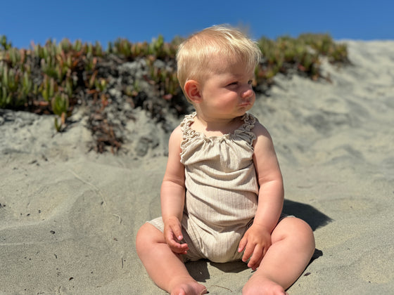Baby girl sitting in the sand while wearing oatmeal colored ruffled baby romper 