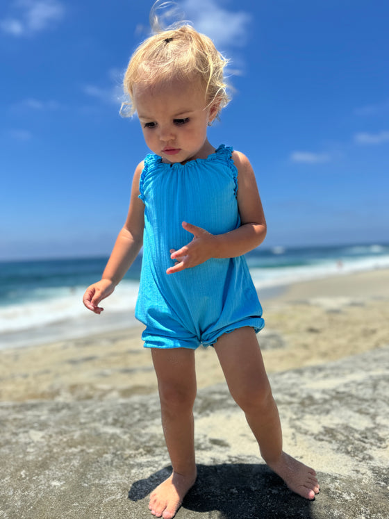 a baby girl model at the beach wearing a bright blue romper
