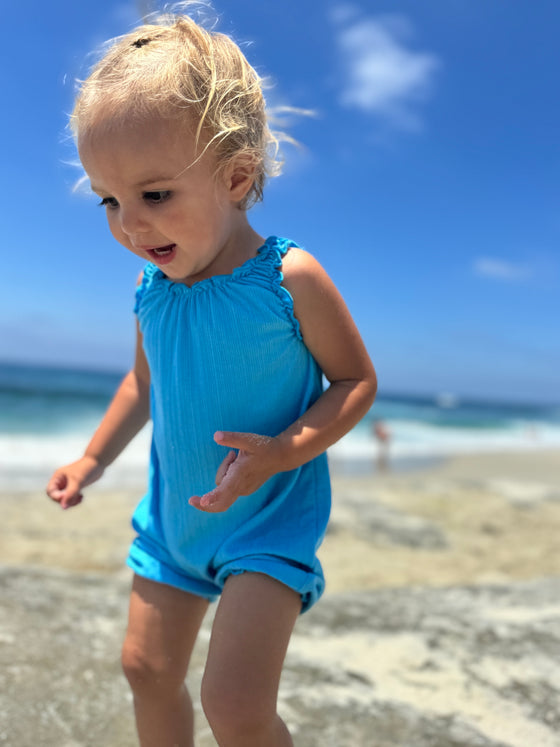 Smiling baby girl model playing on the beach while wearing a bright blue romper