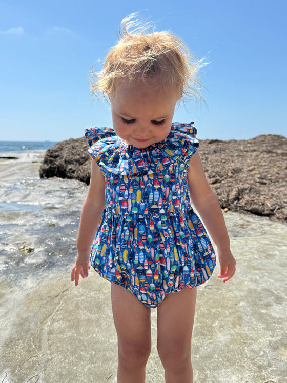 Toddler wearing Vignette’s Maya bubble romper in nautical buoy print, enjoying a sunny day at the beach. This stylish and comfortable baby outfit is perfect for seaside adventures and summer fun. Shop this adorable coastal-inspired bubble for your little one’s wardrobe.”