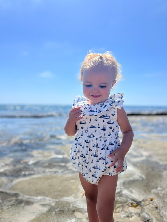 At the beach  a toddler smiles wide ad she wears Vignette’s Maya romper with its nautical boat print, creating a picture-perfect blend of comfy style and sunny beach vibes.