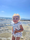 A toddler enjoys a sunny day on the beach wearing Vignette’s Maya romper in nautical boats, a perfect choice for seaside adventures and warm weather fun.
