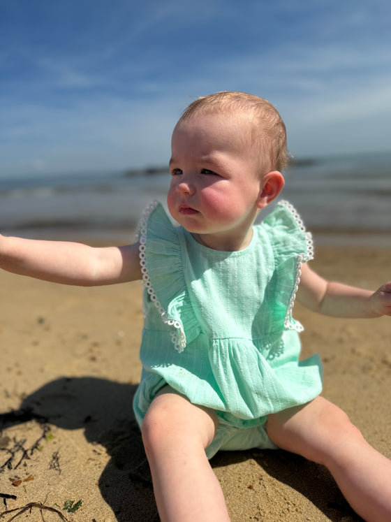 A baby girl sits in the soft sand at a Southern California beach, wearing the Vignette Girls Poppy Bubble in aqua. The romper’s playful ruffle flutter sleeves flutter gently in the breeze, making it the perfect outfit for a sunny day by the ocean.
