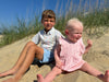 A baby girl enjoys a sunny day on the beach with her big brother. she is wearing the Poppy Bubble in pink, complete with dainty ruffle sleeves that add a sweet touch to her outfit. Her big brother, in complementary shades wearing a nice preppy shirt and shorts  from Me and Henry Boys,
