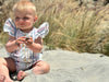A blonde baby girl enjoys a day at the beach, dressed in the Cammie Bubble in Pink and Blue Multi Stripe. The flutter sleeves flutter gently in the ocean breeze, while the multi-stripe pattern adds a playful and vibrant touch to her beachside look. This adorable baby bubble romper is perfect for keeping your little one comfortable and stylish in the sun.