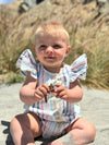 On a sunny beach, a blonde toddler looks adorable in the Cammie Bubble in Pink and Blue Multi Stripe. The soft, lightweight fabric and flutter sleeves make this romper an ideal choice for a warm day at the beach. The pink and blue stripes add a fresh and fun vibe, perfect for a relaxed, stylish day by the ocean.