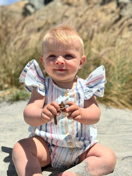 On a sunny beach, a blonde toddler looks adorable in the Cammie Bubble in Pink and Blue Multi Stripe. The soft, lightweight fabric and flutter sleeves make this romper an ideal choice for a warm day at the beach. The pink and blue stripes add a fresh and fun vibe, perfect for a relaxed, stylish day by the ocean.