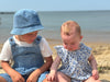 Brother and sister pair at the beach wearing denim overalls and a bucket hat and a baby wearing Light blue floral print baby bubble 