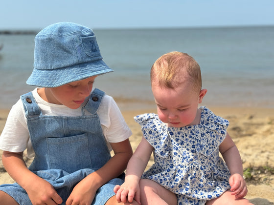 Brother and sister pair at the beach wearing denim overalls and a bucket hat and a baby wearing Light blue floral print baby bubble 