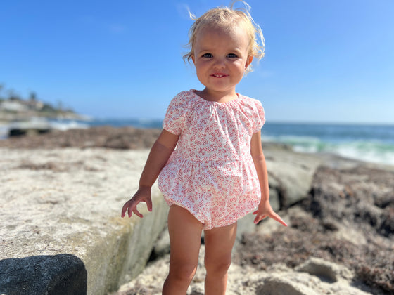 A baby playing at the beach wearing a white baby bubble with delicate coral floral pattern