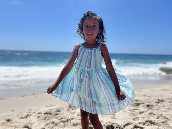 A child model at the beach wearing a blue striped sun dress with a full skirt 