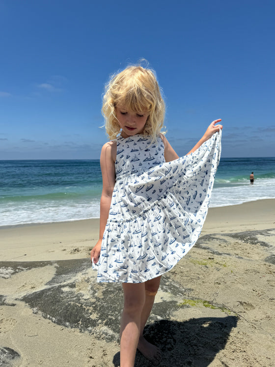 A smiling girl looking like a ballerina while playing at the beach in a white sun dress with all over nautical boat print in blue
