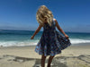 a child looking down at her feet with the ocean behind her  while wearing a blue Dress featuring an all over nautical buoy print