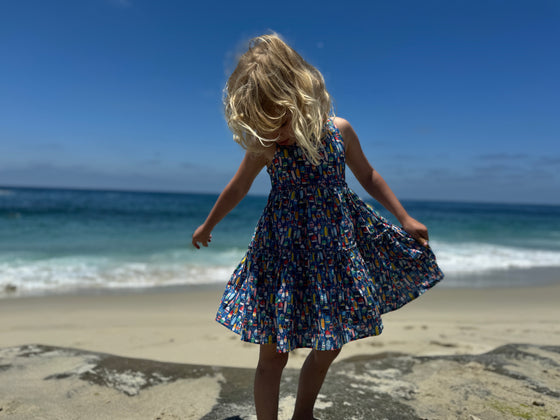 a child looking down at her feet with the ocean behind her  while wearing a blue Dress featuring an all over nautical buoy print