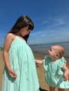 In a lively beach moment, a baby in the Vignette Girls Poppy Bubble in aqua looks lovingly at her big sister, who wears a matching dress. The two share an affectionate gaze while enjoying a day by the ocean, showcasing the perfect sibling bond in coordinating outfits.