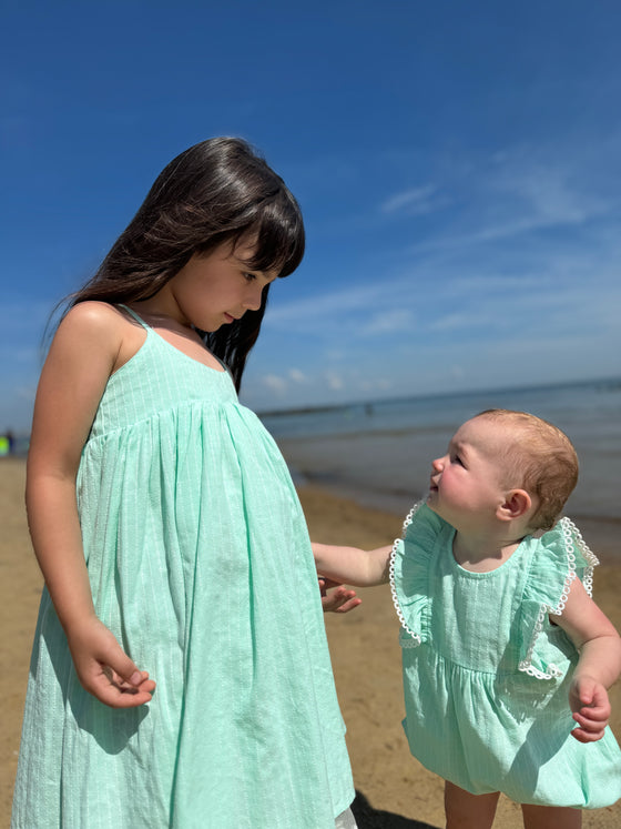 In a lively beach moment, a baby in the Vignette Girls Poppy Bubble in aqua looks lovingly at her big sister, who wears a matching dress. The two share an affectionate gaze while enjoying a day by the ocean, showcasing the perfect sibling bond in coordinating outfits.