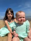 A baby girl wears the Vignette Girls Poppy Bubble in aqua while seated with her big sister, who is dressed in a complementary shade. The two siblings share a moment of sweetness, with their matching outfits creating a coordinated, stylish look.
