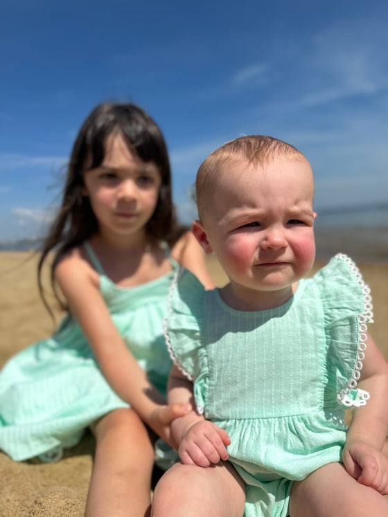 A baby girl wears the Vignette Girls Poppy Bubble in aqua while seated with her big sister, who is dressed in a complementary shade. The two siblings share a moment of sweetness, with their matching outfits creating a coordinated, stylish look.

