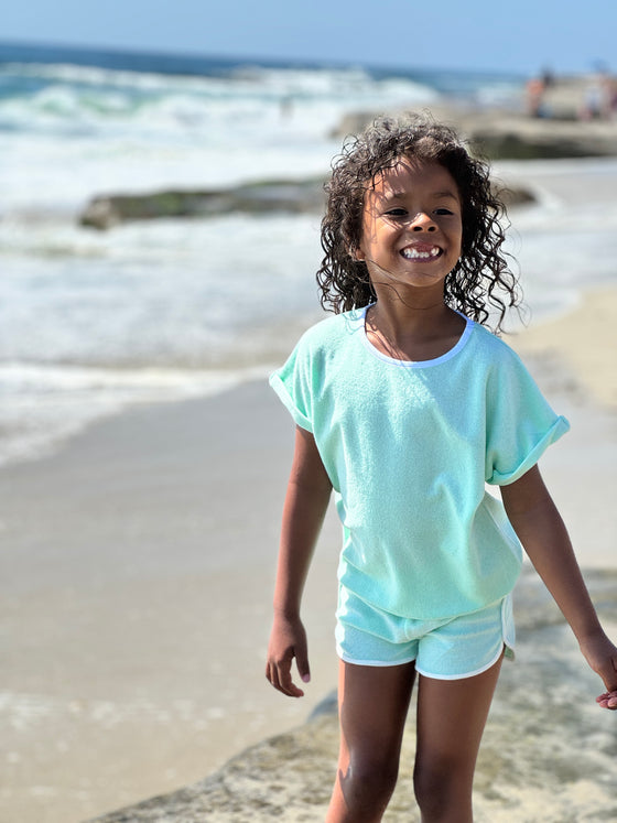 Youg girl smiling at beach wearing seafoam green Terry cloth shirt and matching shorts