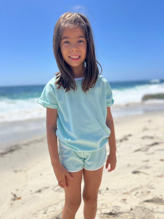 child model at the beach wearing a mint colored Terry cloth shirt and matching shorts